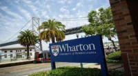 Wharton Edge at UPenn showcased through the Wharton School sign at the San Francisco campus, with palm trees, Bay Bridge, and a light rail train in the background.