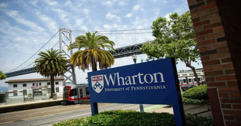 Wharton Edge at UPenn showcased through the Wharton School sign at the San Francisco campus, with palm trees, Bay Bridge, and a light rail train in the background.