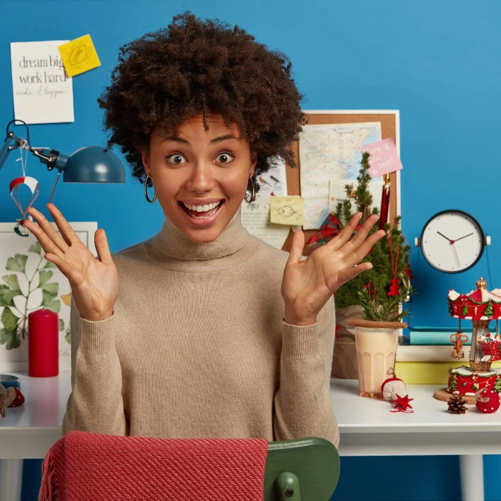 joyful upbeat dark skinned woman has both palms raised sits desktop with other holiday attributes