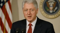 Bill Clinton speaking at a podium with the U.S. flag and presidential seal in the background.