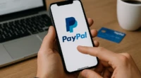 Person using a smartphone with the PayPal app open beside a laptop and coffee cup.